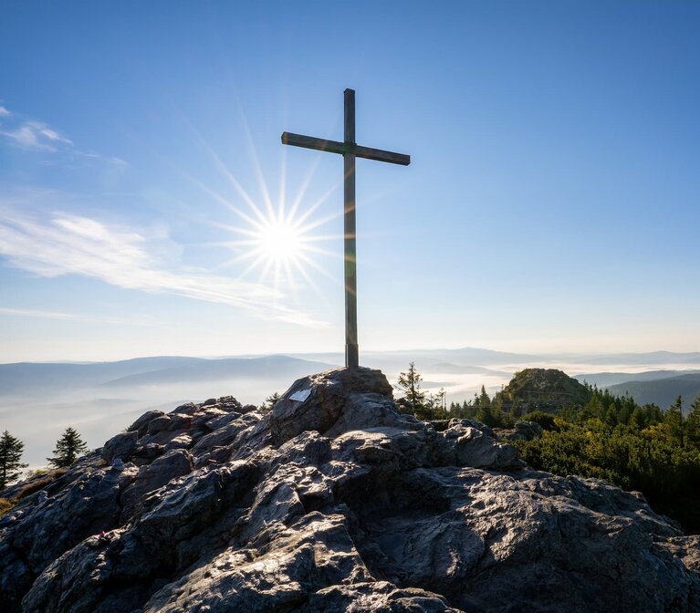Der Blick fällt auf das Gipfelkreuz des Großen Arbers. Von hinten scheint die Sonne das Kreuz an. Im Tal liegen Wolken, aber darüber ist der Himmel blau. | © Bodenmais Tourismus & Marketing GmbH