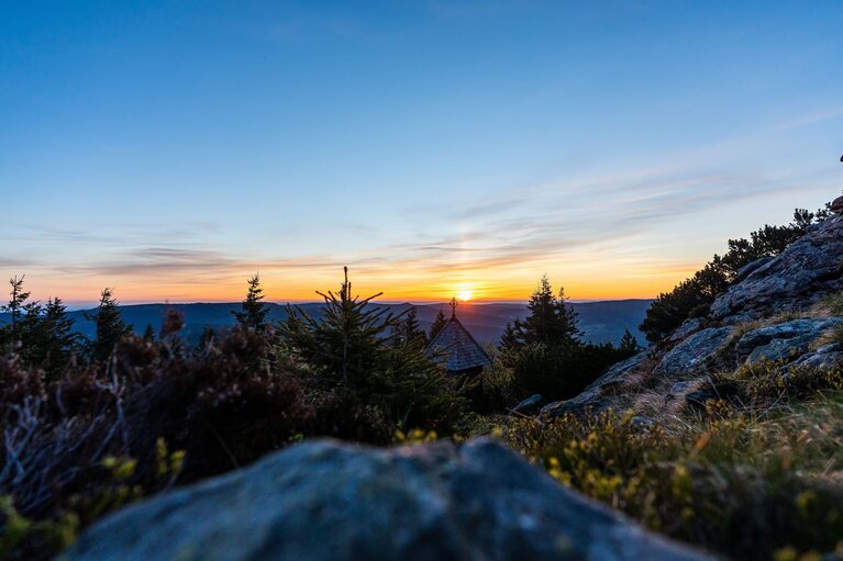 Über einen Stein und Sträucher hinweg erblickt man den Sonnenuntergang hinter der Arberkapelle. | © Bodenmais Tourismus & Marketing GmbH