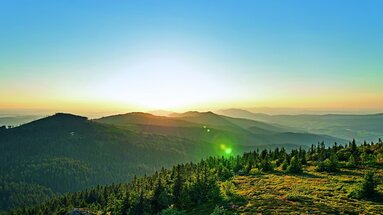 Vom großen Arber aus blickt man auf die Berge des Bayerischen Waldes. Der Himmel ist blau und die Sonne scheint grell, doch der Horizont verfärbt sich schon orange. | © Bodenmais Tourismus & Marketing GmbH
