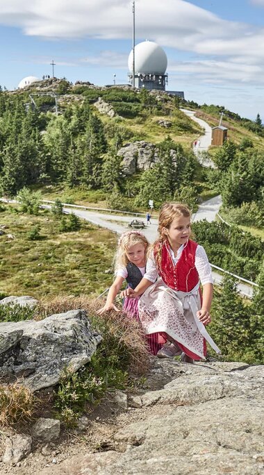 Zwei Mädchen in Dirndl klettern auf den Nebengipfel des Großen Arbers. Im Hintergrund ragt einer der beiden Radartürme in die Höhe. | © Bodenmais Tourismus & Marketing GmbH
