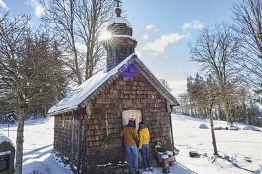Eine Frau und ein Mann schauen durch die Tür der Kapelle am Harlachberg. Es liegt Schnee der durch die Sonne glitzert. | © Bodenmais Tourismus & Marketing GmbH
