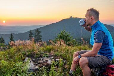 Ein Mann sitzt im Sommer bei Sonnenaufgang auf einem Fels am Berg und trinkt aus einer Tasse. | © Bodenmais Tourismus & Marketing GmbH