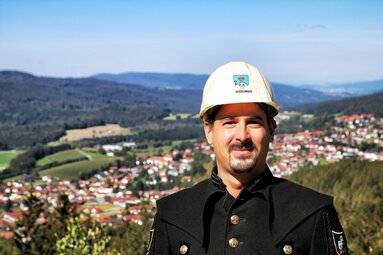 Ein Mann mit schwarzer Uniform und weißem Helm grinst in die Kamera. | © Historisches Besucherbergwerk Bodenmais BHS GmbH