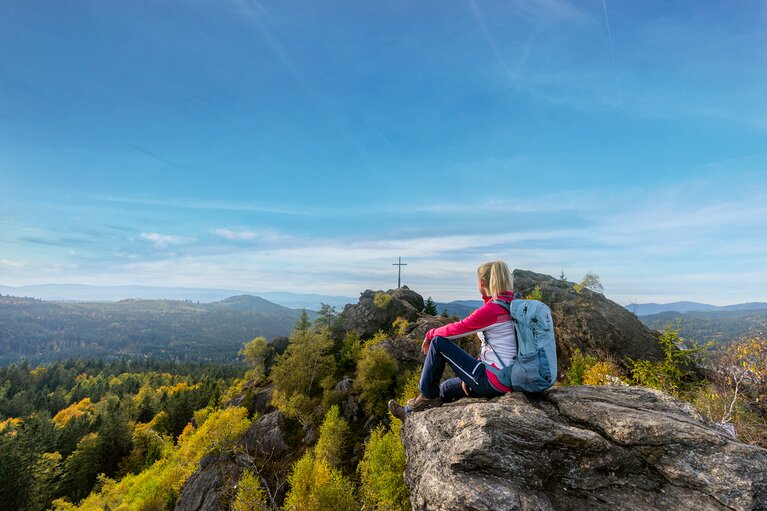 Wanderin mit Rucksack auf einem Felsen sitzend, blickt auf den Silberberg Gipfel mit Gipfelkreuz. | © Bodenmais Tourismus & Marketing GmbH