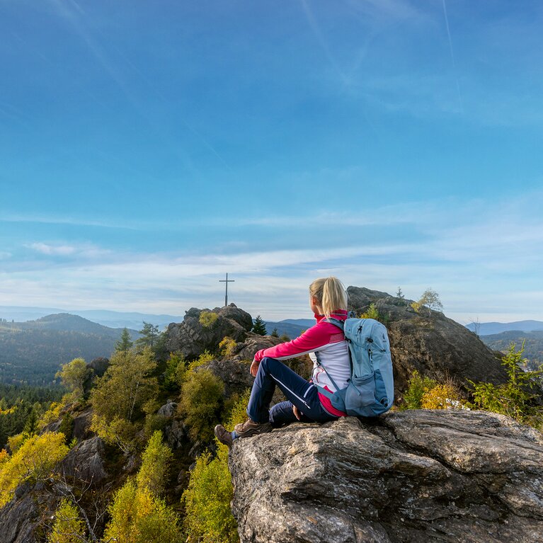 Wanderin mit Rucksack auf einem Felsen sitzend, blickt auf den Silberberg Gipfel mit Gipfelkreuz. | © Bodenmais Tourismus & Marketing GmbH