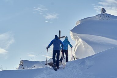 Zwei Wanderer mit Schneeschuhen und Stöcken wandern auf ein Gipfelkreuz zu. Alles ist mit Schnee bedeckt und rechts vom Weg haben sich große Schneeformationen gebildet. | © Bodenmais Tourismus & Marketing GmbH