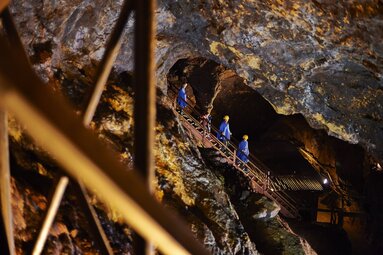 Eine Besuchergruppe mit blauen Kitteln und gelben Helmen geht im Bergwerk des Silberbergs eine Treppe hinunter | © Bodenmais Tourismus & Marketing GmbH