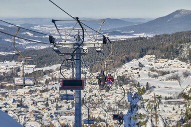 Mit dem Skilift fahren mehrere Skifahren den Silberberg hinauf. Im Hintergrund sieht man das verschneite aber sonnige Bodenmais. | © Bodenmais Tourismus & Marketing GmbH