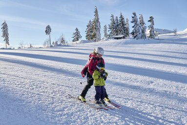 Eine Mutter bringt ihrem Kind das Skifahren bei, indem sie es vor sich hält. Auf dem Rücken trägt sie noch ein zweites Kind. | © Bodenmais Tourismus & Marketing GmbH