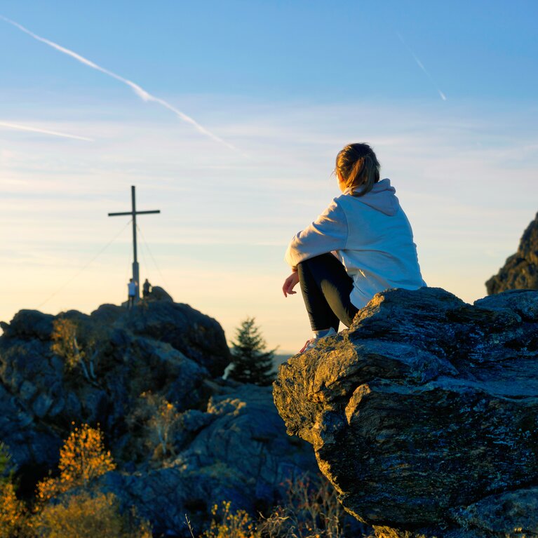 Eine Frau sitzt mit dem Rücken zur Kamera auf einem Feld. Sie blickt in Richtung des Gipfelkreuzes vom Silberberg. | © Bodenmais Tourismus & Marketing GmbH
