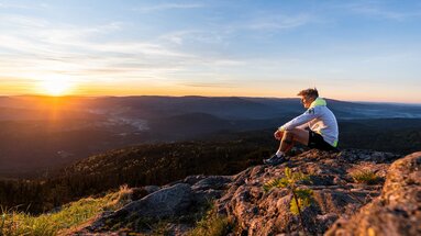 Ein Mann sitzt in Sportkleidung am großen Arber und genießt den Sonnenaufgang, welcher alles in warmes Licht hüllt. | © Bodenmais Tourismus & Marketing GmbH