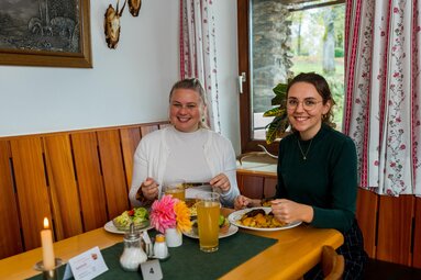 Zwei Frauen sitzen in einer urigen Wirtschaft. Am Tische stehen Getränke und deftige Hauptspeisen sowie Beilagensalate. | © Bodenmais Tourismus & Marketing GmbH