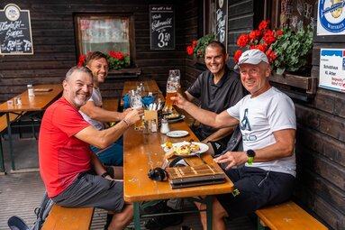 Vier Männer sitzen an einem Biertisch. Sie stoßen mit Weißbier an. Am Tisch steht Essen. | © Bodenmais Tourismus & Marketing GmbH