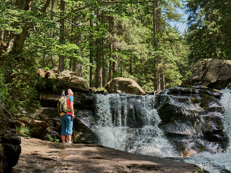 Eine Wanderin mit Rucksack blickt auf die Risslochwasserfälle umgeben von Bäumen. | © Bodenmais Tourismus & Marketing GmbH