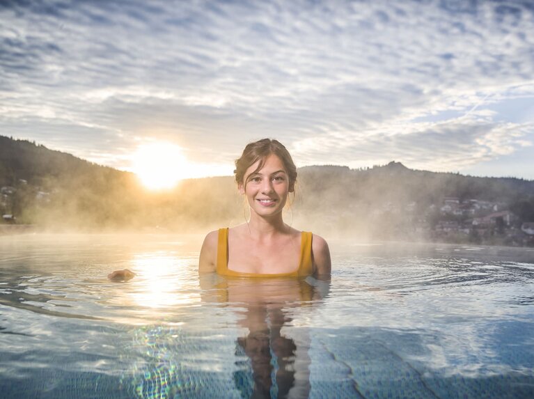Eine Frau ist bis zu den Schultern in einem Pool. Die Haare hat sie nach oben gebunden und sie lacht in die Kamera. Hinter ihr ist Bodenmais sowie der Silberberg und der Sonnenuntergang zu erkennen. | © Bodenmais Tourismus & Marketing GmbH