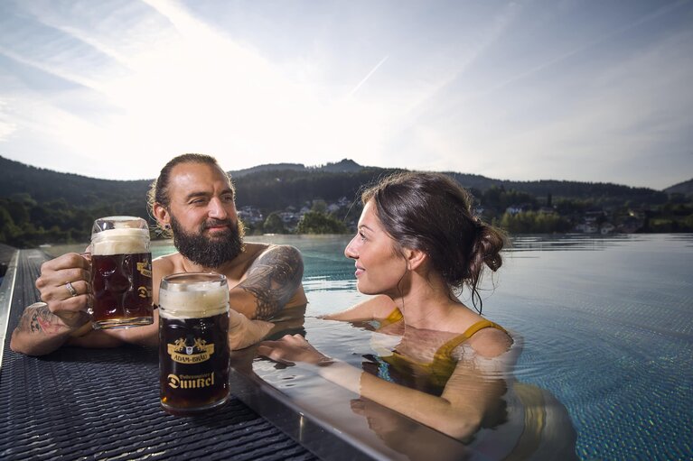 Eine Frau und ein Mann sind im Pool und lehne am Rand. Sie lächeln sich an und haben dabei jeder einen Maßkrug mit dunklem Bier in der Hand, mit dem sie anstoßen. | © Bodenmais Tourismus & Marketing GmbH