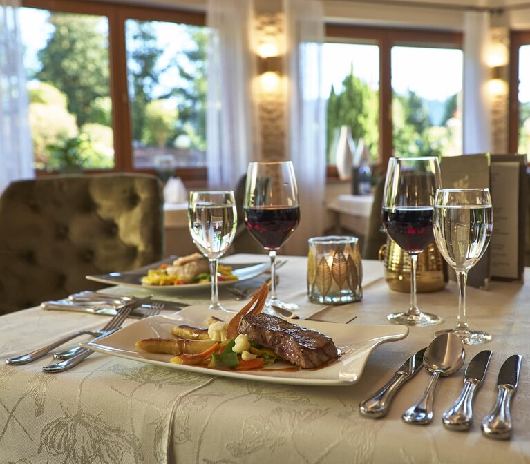 Auf einem Tisch mit weißer Tischdecke ist für zwei Personen und vier Gänge gedeckt. Auf einem Teller ist ein saftiges Steak mit Gemüse angerichtet. Daneben steht ein Glas Rotwein | © Bodenmais Tourismus & Marketing GmbH