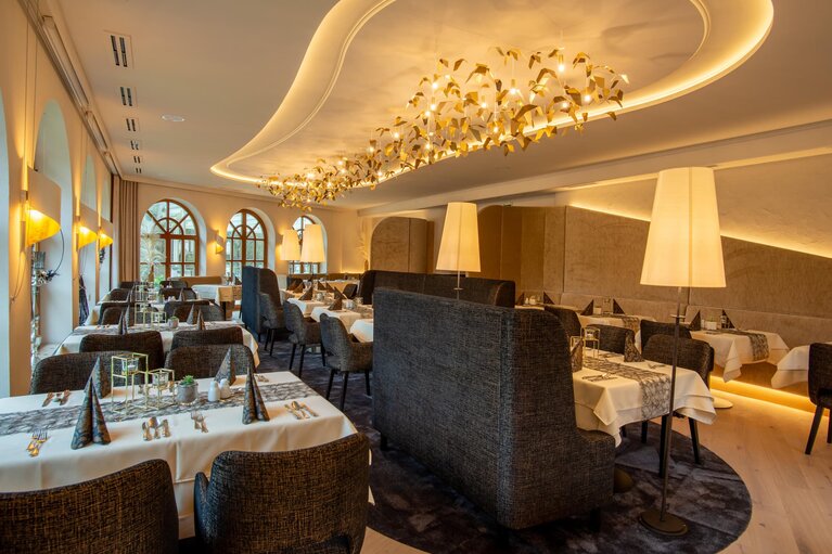 Das Restaurant des Hotels Hammerhof ist in den Farben Weiß, Grau, Beige und Gold gehalten. Die Tische sind mit je vier gepolsterten Stühlen ausgestattet. Goldene Lampen an der Wand als auch eine sehr auffällige goldene Lampe an der Decke sorgen für stimmungsvolles Licht. | © Bodenmais Tourismus & Marketing GmbH