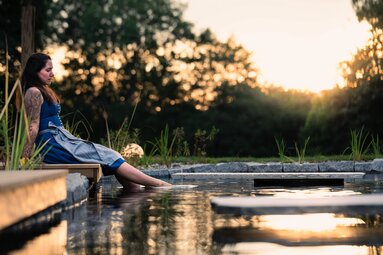 Eine Frau in Tracht lässt ihre Beine in einem Naturbadeteich baumeln. Hinter dem Pool ist eine Wiese mit angrenzender Hecke, hinter der die Sonne verschwindet. | © FOUQUET PRODUCTION