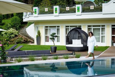 Eine Frau in Bademantel geht am Außenpool des Hotels Hammerhof entlang. Neben dem Pool befindet sich ein Sonnendeck sowie eine Wiese mit verschiedenen Sitz- und Liegemöglichkeiten. | © Bodenmais Tourismus & Marketing GmbH