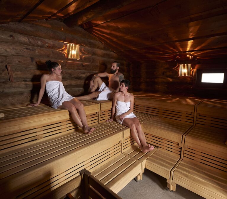 Ein Mann und zwei Frauen sitzen mit umgebundenen Handtüchern zusammen in der Sauna. | © Bodenmais Tourismus & Marketing GmbH