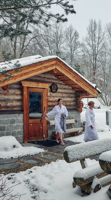 Zwei Frauen in weißem Bademantel gehen aus einer Blocksaune in den verschneiten Saunagarten. | © Bodenmais Tourismus & Marketing GmbH