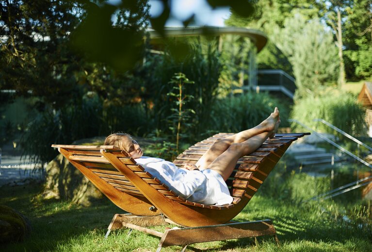 Eine Frau liegt mit Bademantel und geschlossenen Augen auf einer ergonomisch geformten Holzliege im Saunagarten. | © Bodenmais Tourismus & Marketing GmbH