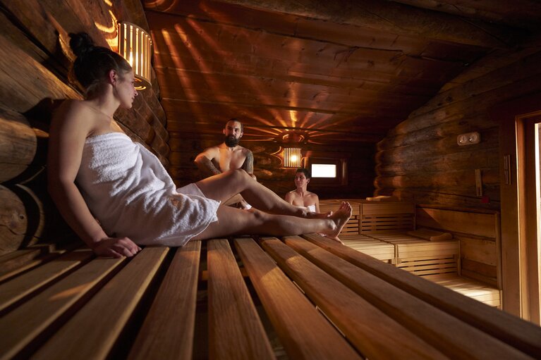 Zwei Frauen und ein Mann, mit Handtüchern bekleidet. sitzen zusammen in der Sauna | © Bodenmais Tourismus & Marketing GmbH