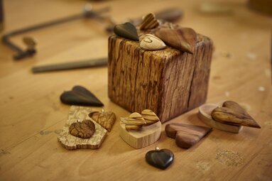 Auf einem Tisch und einem kleinen Holzklotz liegen verschiede Schmuckstücke aus Holz. Darunter Herzohrringe mit verschiedenen Marmorierung oder Herz-Anhänger | © Bodenmais Tourismus & Marketing GmbH