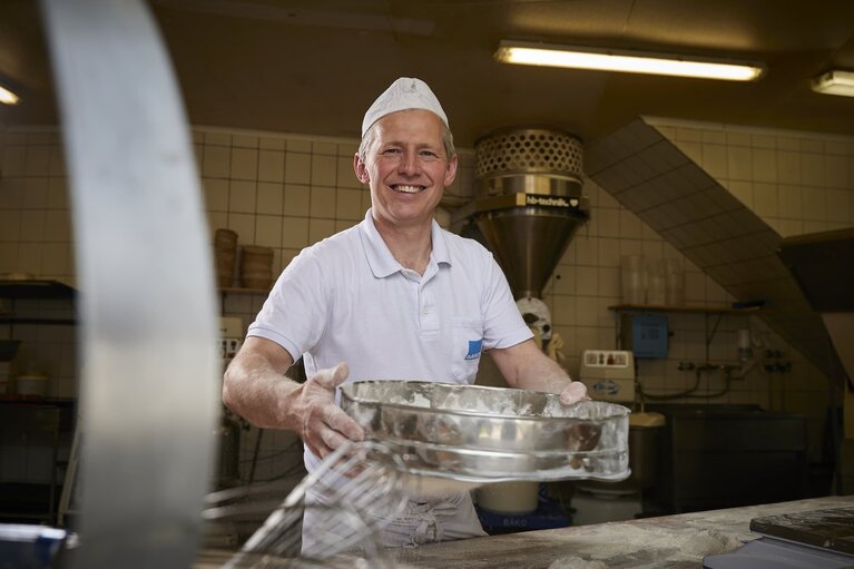 Ein lächelnder Bäcker steht in seiner Backstube und hält einen großen Sieb vor sich. | © Bodenmais Tourismus & Marketing GmbH