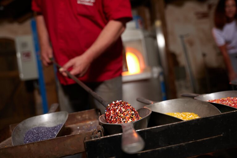 Ein Glasbläser hält heißes, zähflüssiges Glas in farbiges Granulat. Hinter ihm brennt der Ofen. | © Bodenmais Tourismus & Marketing GmbH