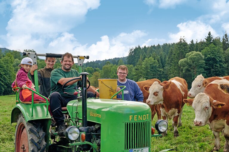 Auf einer Weide steht ein grüner Traktor. Darauf sitzt ein lachender Mann und zwei Kinder. Daneben steht ein weiterer Mann und Kühe. | © Bodenmais Tourismus & Marketing GmbH